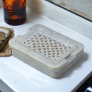 May include: A light gray soap dish made of stone with a decorative, carved, openwork design. The dish is rectangular and has a raised edge.