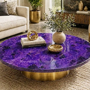 May include: A circular coffee table with a striking purple surface and a gold-coloured base. The table has a polished, gem-like appearance. Decorative items, including books and a gold bowl, are arranged on the table's surface. The table is in a living room setting.