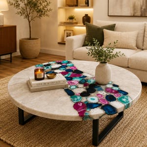 Puede incluir: Mesa de centro redonda con tapa de mármol blanco e incrustaciones de ágata de colores. Las patas son de metal negro. Sobre la mesa hay una vela encendida y libros. La mesa tiene un diámetro de aprox. 120 cm.
