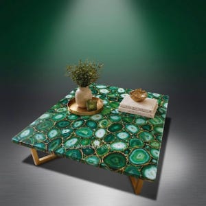May include: A square coffee table with a green agate stone top, edged with gold resin. The table has a natural stone pattern and is supported by light-coloured wooden legs. A vase, tray, and books are on the table.