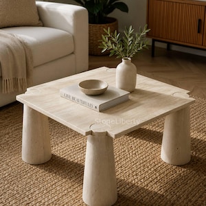 May include: A square, beige stone coffee table with four cylindrical legs. The table has a light-colored surface with a book, bowl, and vase with greenery. The text "LIVE BEAUTIFULLY" is visible on the book.