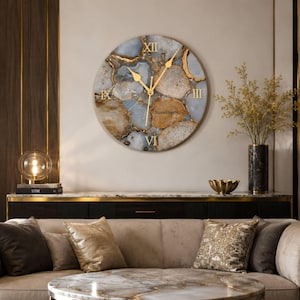 May include: A round wall clock featuring a natural agate stone pattern in shades of gray, white, and brown with gold-colored veins. The clock face displays gold Roman numerals at the twelve, three, six, and nine positions and the text Stoneliberry.