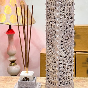 May include: A set of two carved stone incense holders. The larger holder is a tall, rectangular tower with an intricate floral design. The smaller holder is a square base with a round top and a small hole in the center. Both holders are made of a light gray stone.