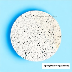 Op de afbeelding: Een ronde, witte terrazzo-onderzetter met zwarte en groene vlekken. De onderzetter bevindt zich op een lichtblauwe achtergrond. EpoxyMarbleAgateShop