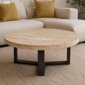 May include: A round coffee table featuring a beige, porous travertine stone top with a natural textured surface and the text StoneLiberty printed on it. The table is supported by a black metal base with three legs connected in a central cross design.