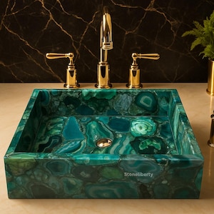 May include: A square, green stone sink with a gold faucet and handles. The sink is made of polished, irregularly shaped pieces of green and teal stone. The faucet and handles are gold-colored. The sink is labeled "Stoneliberty".