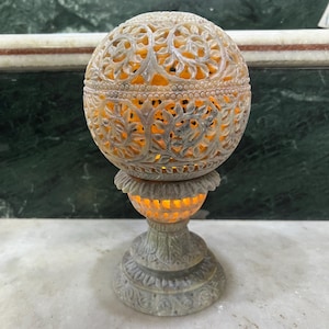 May include: A carved stone sphere with intricate floral designs sits on a matching base. The sphere is illuminated from within, casting a warm glow.