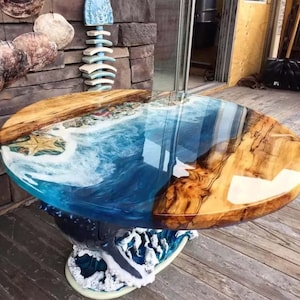 Op de afbeelding: Een ronde houten tafel met een blauw en wit epoxyhars blad dat lijkt op een oceaan golf. De tafelpoot is gebeeldhouwd om te lijken op een oceaan golf.