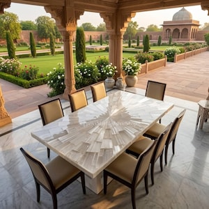 Pode incluir: Uma mesa de jantar retangular com um tampo branco com padrão de estrela e oito cadeiras bege. A mesa está montada num pátio de mármore, com um jardim e um edifício ao fundo. A mesa tem aproximadamente 183 cm de comprimento.