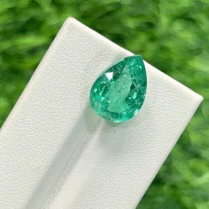 Peut inclure: Une émeraude verte en forme de poire. La pierre précieuse est d'un vert vif et repose sur une surface blanche, avec un arrière-plan vert flou. L'émeraude présente des inclusions internes et des facettes visibles.