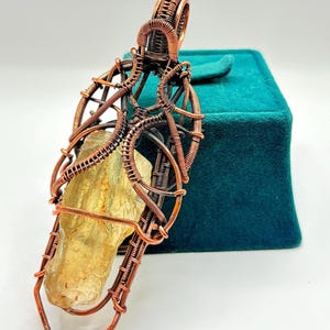 May include: A handcrafted pendant featuring a raw, golden-yellow crystal encased in intricate copper wire. The wire is meticulously woven and shaped, creating a cage-like design around the stone. The pendant is displayed against a teal background.