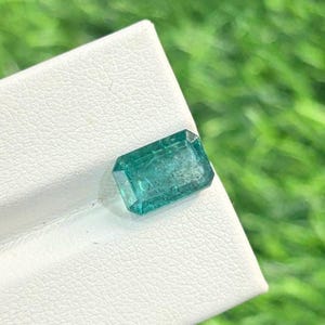 Könnte beinhalten: Ein rechteckiger, smaragdgeschliffener Edelstein in tiefem Teal. Der Edelstein wird auf einer weißen Oberfläche vor einem verschwommenen grünen Hintergrund präsentiert, was auf eine natürliche Umgebung hindeutet. Die Facetten des Steins fangen das Licht ein und zeigen seine Klarheit und Brillanz.