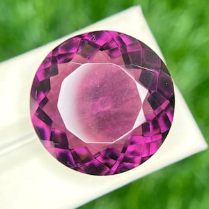 May include: A faceted, round gemstone with a deep magenta hue. The gem is set against a white surface, with a blurred green background. The cut of the stone creates multiple facets, reflecting light.