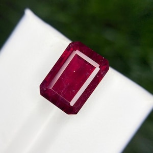 May include: A rectangular, faceted ruby gemstone with a deep red hue. The gem has a clear, rectangular interior and a white border. The gemstone is set against a white background with a blurred green backdrop.