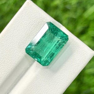 Puede incluir: Una esmeralda verde vibrante, de corte rectangular. La gema está sobre una superficie blanca, con un fondo verde borroso. Las facetas de la esmeralda reflejan la luz, mostrando su claridad y color. Ideal para joyería.