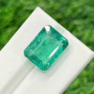Op de afbeelding: Een rechthoekige smaragd met een levendige groene kleur. De edelsteen ligt op een wit oppervlak, met een vage groene achtergrond. De facetten van de smaragd reflecteren het licht, wat de helderheid en glans laat zien. Een close-up benadrukt het gepolijste oppervlak van de edelsteen.
