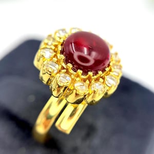 Puede incluir: Un anillo de oro con una gran piedra preciosa ovalada de color rojo oscuro en el centro, rodeada de piedras preciosas más pequeñas y claras. La banda del anillo está dividida y se curva hacia arriba, creando un diseño elegante. El anillo está sobre un fondo oscuro.