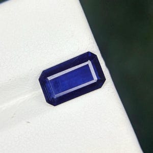 May include: A rectangular, deep blue gemstone with a white interior, set against a white surface. The gemstone has a faceted cut, reflecting light and showcasing its vibrant colour. The gem is likely a sapphire or similar precious stone.