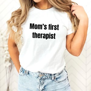May include: White t-shirt with the text "Mom's first therapist" in black, bold letters. The shirt is a classic crew neck style.
