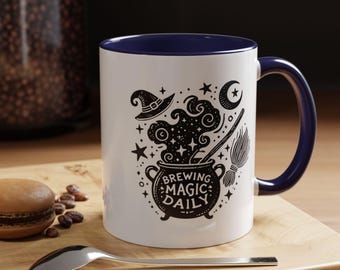Puede incluir: Taza de cerámica blanca con borde y asa azules. La taza presenta una ilustración en blanco y negro de un caldero con vapor, un sombrero de bruja, una escoba y estrellas. El texto "Brewing Magic Daily" está impreso en el caldero.