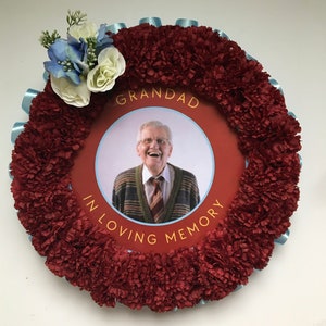 May include: A red and blue floral wreath with a photo of a man smiling. The wreath has a white circle with the text "Grandad In Loving Memory".