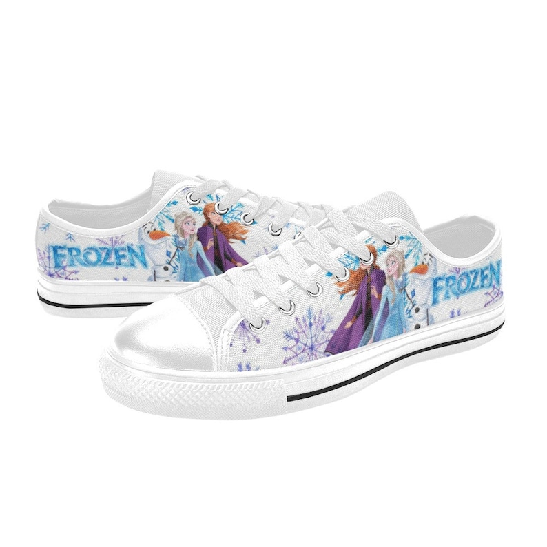May include: White canvas low-top sneakers with a Frozen theme. The shoes feature a white background with a blue and purple snowflake pattern. The characters Elsa and Anna from the movie Frozen are featured on the shoes, along with Olaf the snowman. The words "Frozen" are printed on the side of the shoes.
