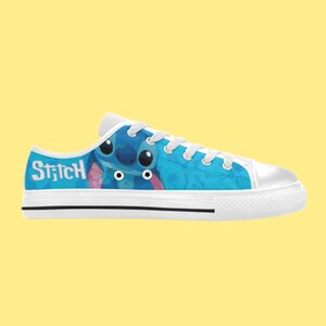 May include: White canvas low-top trainers with a blue and white design featuring Stitch from Lilo & Stitch. The text "Stitch" is printed on the side of the shoe.