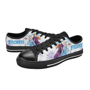 May include: Black canvas low-top sneakers with a white background featuring a Frozen design with Elsa and Anna. The shoes have black laces and a white stripe around the sole. The words "Frozen" are printed on the side of the shoes.