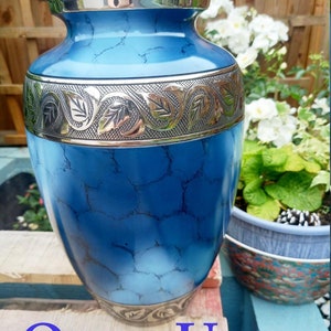 May include: A blue urn with a silver lid and decorative band. The urn has a marbled effect and the text "Quest Urns" is at the bottom. The urn is displayed outdoors with plants in the background.