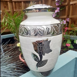 May include: White ceramic urn with a silver floral design. The urn has a silver lid and a silver band around the middle. The urn is sitting on a blue surface. Quest Urns.