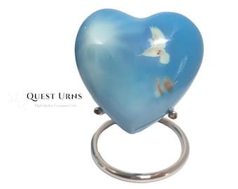 Puede incluir: Una pequeña urna de cremación en forma de corazón azul con un diseño de paloma blanca y nubes. La urna está sobre un soporte de metal plateado.