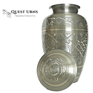 Op de afbeelding: Zilveren crematie urn met een gegraveerd bloemenmotief. De urn heeft een deksel met een bijpassend motief.  "QUEST URNS High Quality Cremation Urns" is afgedrukt op de afbeelding.