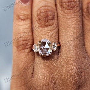 Op de afbeelding: Een ros&eacute;gouden ring met een grote, ovaal geslepen diamant als middensteen, geflankeerd door twee kleinere, ronde diamanten. De ring wordt getoond op een hand met een lichte huidskleur, waardoor de schittering van de edelstenen wordt benadrukt. Een sieraad.