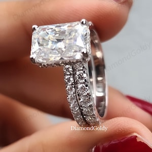 Op de afbeelding: Een rechthoekige diamanten verlovingsring met bijpassende band. De ringen zijn gezet in een zilverkleurig metaal, waarbij de hoofddiamant rechthoekig is. De band is bezet met kleinere diamanten. De afbeelding is goed belicht.