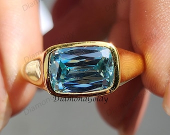 May include: A gold ring with a rectangular, light blue gemstone. The ring has a thick band and a bezel setting that surrounds the gemstone. The gemstone is a light blue color with a rectangular cut. The ring is held by a person.