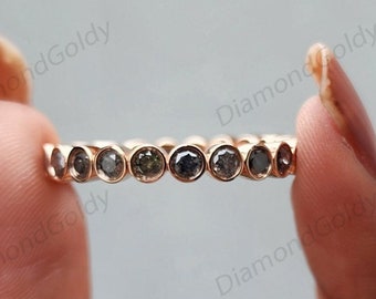 Puede incluir: Un anillo de oro con una serie de diamantes grises redondos engarzados en bisel. El anillo se sostiene entre dos dedos sobre un fondo claro. Una delicada pieza de joyería.
