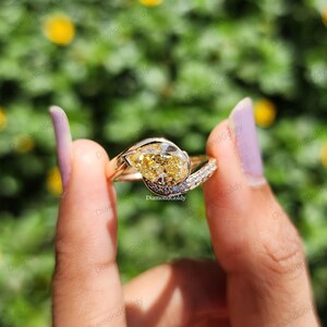 May include: A gold ring with a pear-shaped yellow diamond center stone. The ring has a split shank design with small white diamonds and blue accents. The ring is held up against a blurred green background.