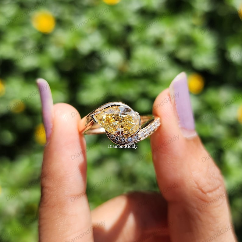 May include: A gold ring with a pear-shaped yellow diamond center stone. The ring has a split shank design with small white diamonds and blue accents. The ring is held up against a blurred green background.