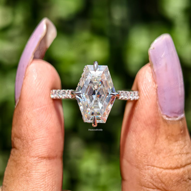 May include: A sparkling engagement ring with a large, geometric-cut diamond as the centerpiece. The ring features a band adorned with smaller, clear stones, creating a brilliant display. The diamond's facets reflect light, showcasing its clarity and brilliance. The ring is held against a blurred green background.