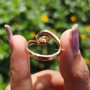 May include: A gold ring with a heart-shaped design, featuring a central, light-colored gemstone. The ring's band curves gracefully, creating a unique and elegant look. The ring is held up against a blurred green and yellow background.