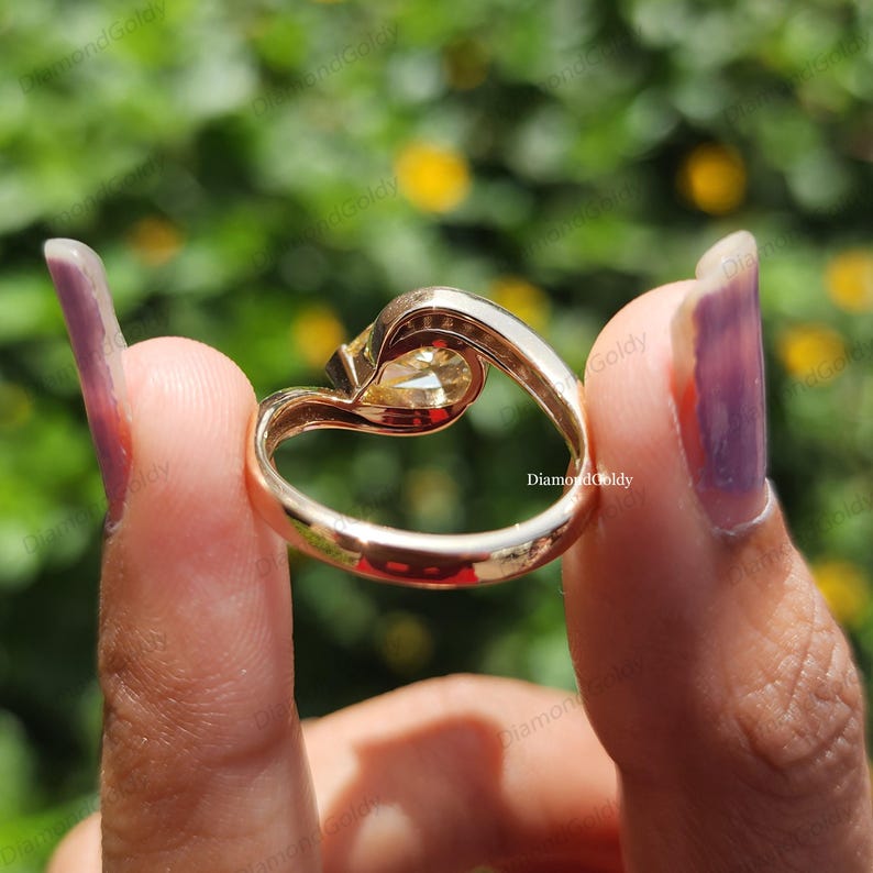 May include: A gold ring with a heart-shaped design, featuring a central, light-colored gemstone. The ring's band curves gracefully, creating a unique and elegant look. The ring is held up against a blurred green and yellow background.