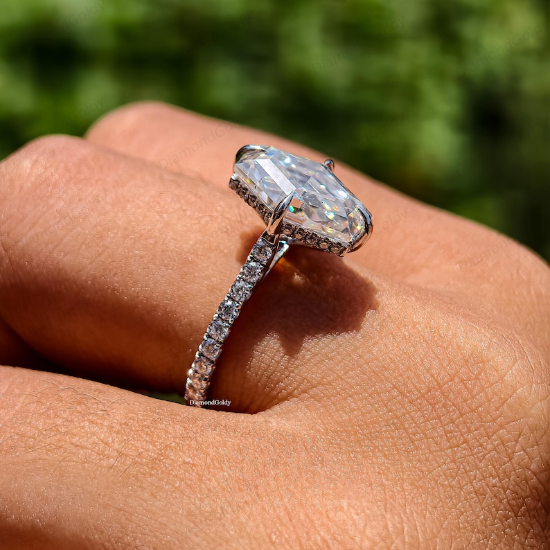 May include: A close-up of a diamond ring with a large, rectangular-cut center stone. The ring has a pave band and a halo of smaller stones around the main diamond. The ring is set in a silver metal.