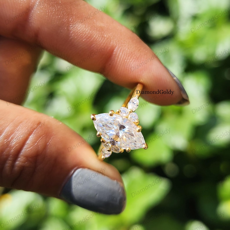 May include: A gold ring featuring a large, marquise-cut diamond as the centerpiece, accented by smaller, round diamonds. The ring is held against a blurred green background, showcasing its sparkle and design. The ring is a piece of jewelry.
