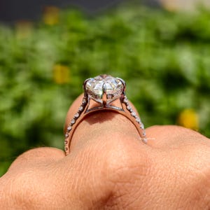 May include: A close-up of a diamond ring with a large, clear center stone. The ring has a silver band with small accent stones. The ring is held up against a blurred green background.