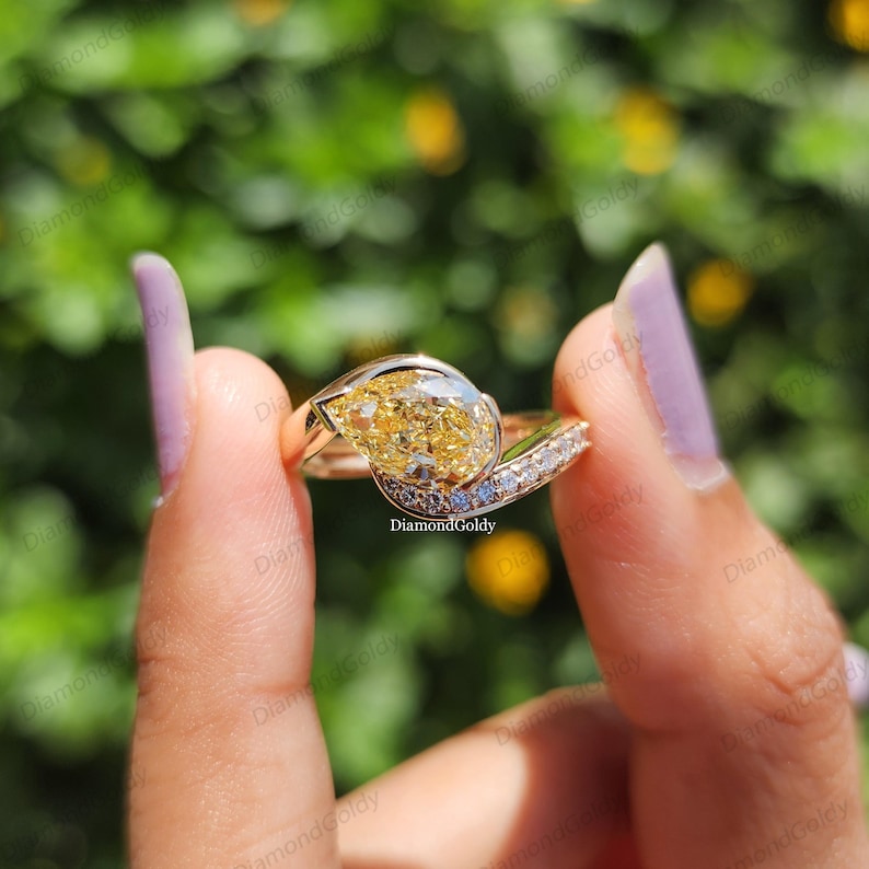 May include: A gold ring featuring a large, pear-shaped yellow diamond as the focal point. The ring has a split shank design, with a row of smaller diamonds set along one side. The ring is held up against a blurred green background.