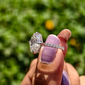 May include: A diamond ring with a large, uniquely cut, clear gemstone. The ring's band is encrusted with small, sparkling diamonds. The ring is held up against a blurred green background, showcasing its brilliance.