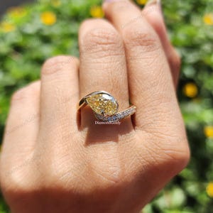 May include: A gold ring featuring a pear-shaped yellow gemstone. The ring has a curved design with a band of small, clear stones. The ring is on a person's finger. The text "Diamond Goldy" is visible.
