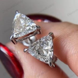 Puede incluir: Un par de pendientes de diamantes. Cada pendiente presenta un gran diamante de talla triangular engastado en un engaste de metal plateado. Los diamantes brillan con facetas internas, reflejando la luz.