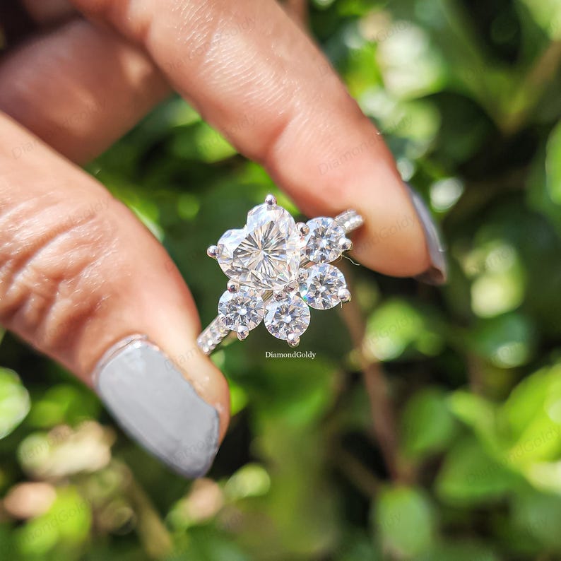 Może przedstawiać: Srebrny pierścionek z diamentem w kształcie serca pośrodku, otoczonym sześcioma okrągłymi diamentami, tworzącymi kwiatowy wz&oacute;r. Pierścionek jest trzymany na tle rozmytego zielonego tła. Widoczny jest tekst "DiamondGoldy".