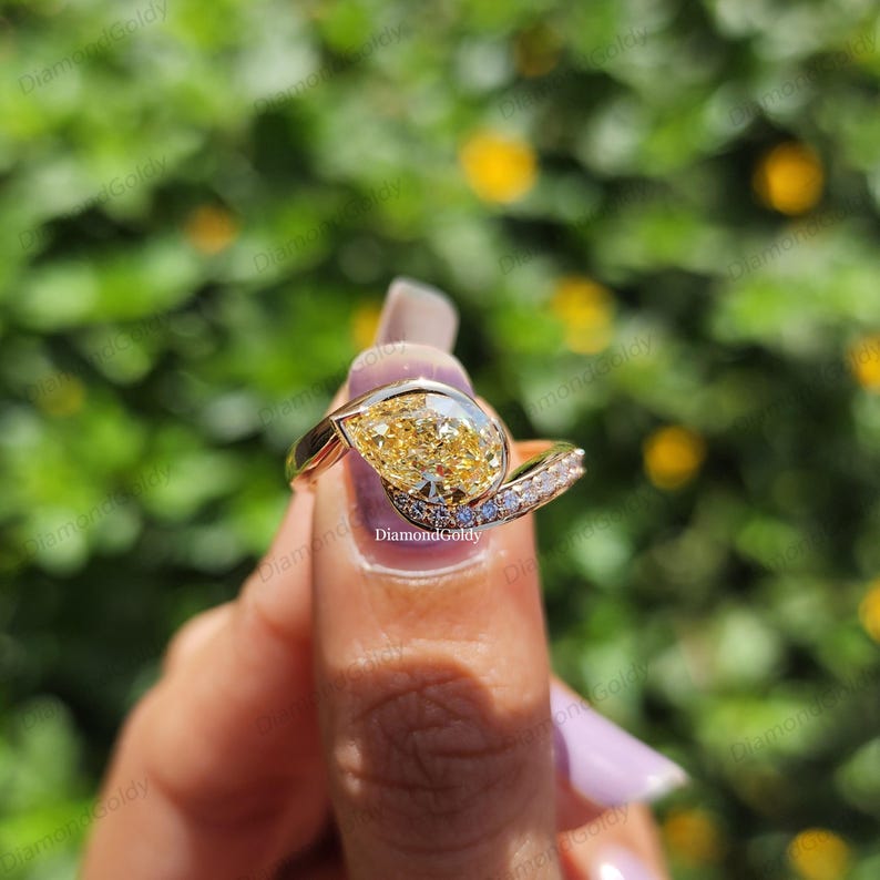 May include: A gold ring featuring a pear-shaped yellow gemstone. The ring has a split shank design with a band of small, clear stones. The ring is held up against a blurred green and yellow background.